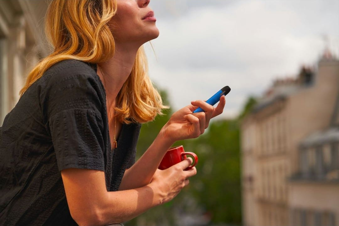 A woman holding a vape and a red coffee cup while relaxing on a balcony. The image represents vaping as a casual and enjoyable activity, aligning with a beginner’s guide to vaping.